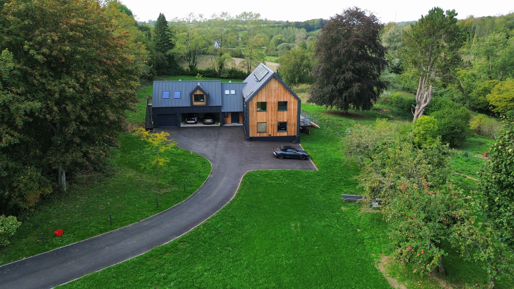 Toft Cottage, Cirencester Modern house surrounded by greenery and trees, with a winding driveway and parked car.